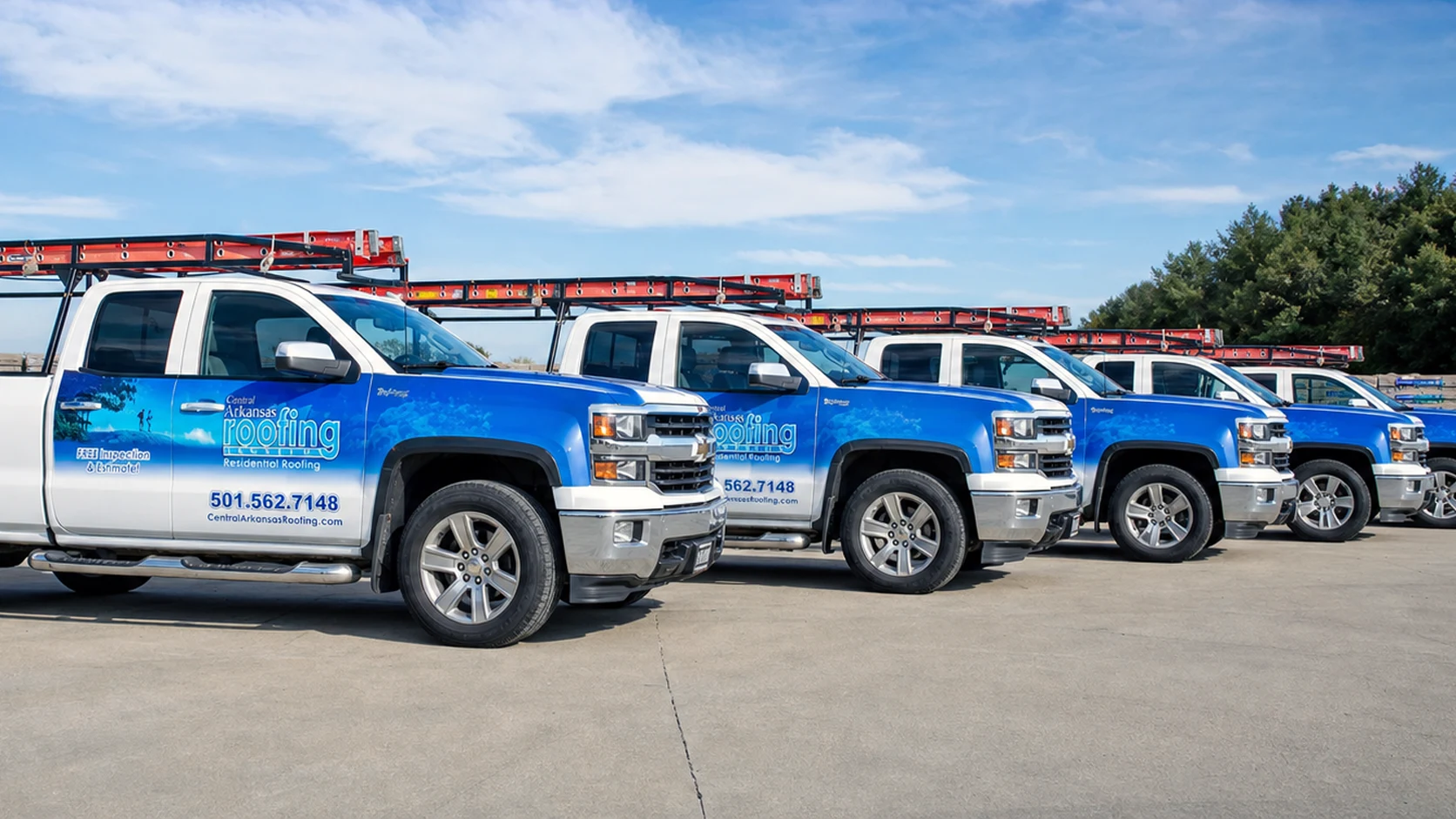 Blue and white Central Arkansas Roofing trucks parked in a row with ladders on their racks